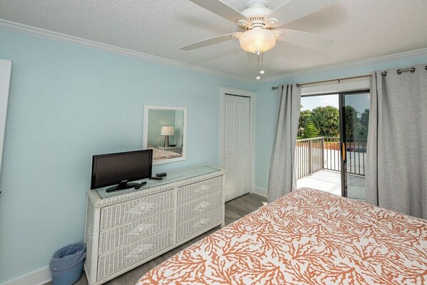 A bedroom with a bed, a television on a dresser, a ceiling fan, and a balcony with curtains.