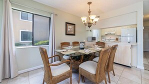 Uma sala de jantar com mesa de vidro, cadeiras de vime, um candelabro e uma cozinha ao fundo.