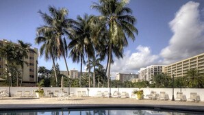Zona junto a la piscina con sillones de descanso y palmeras, con edificios reflejándose al fondo.