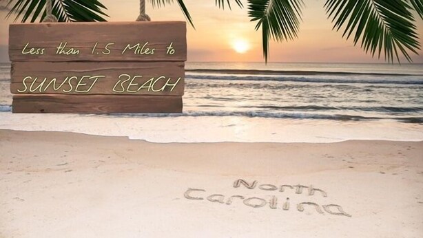 Un panneau en bois indiquant « moins de 2,4 kilomètres jusqu’à Sunset Beach » et « Caroline du Nord », ces inscriptions étant tracées dans le sable.