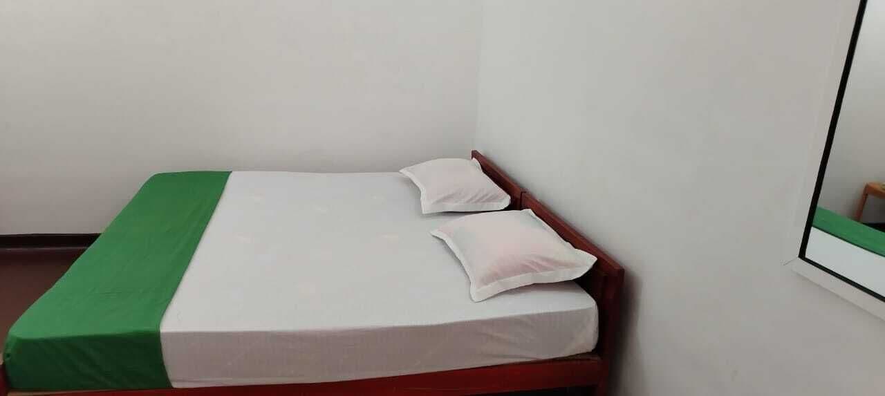 Una habitación pequeña con cama individual, colcha verde y blanca, dos almohadas y cabecera de madera.