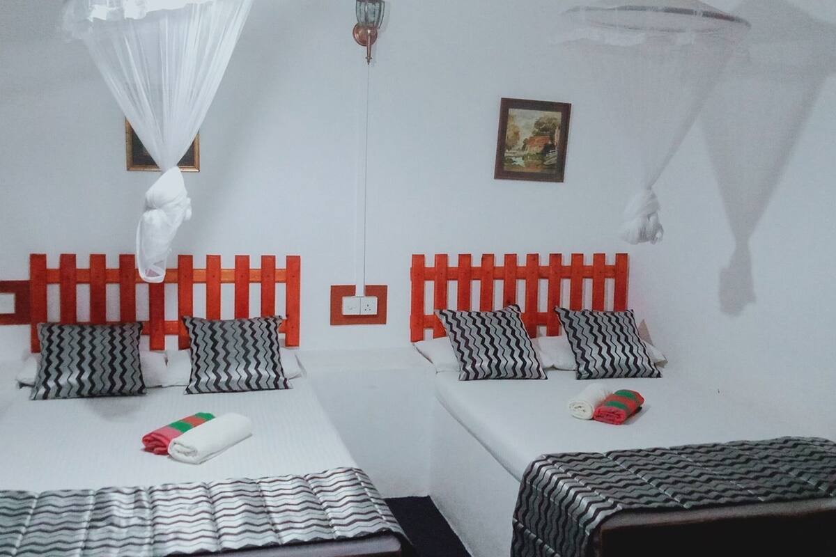 Een kamer met twee bedden, elk met een rode hoofdbord, wit beddengoed en zwart-wit gestreepte kussens. Er staat een klein tafeltje met een geruit tafelkleed en een opgerolde handdoek.