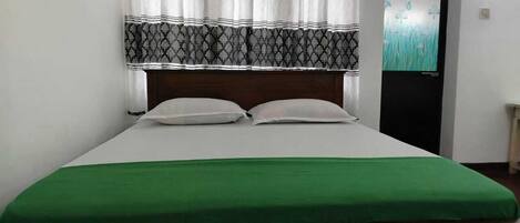 A bed with a green bedspread and white pillows, a patterned headboard, and a window with curtains.