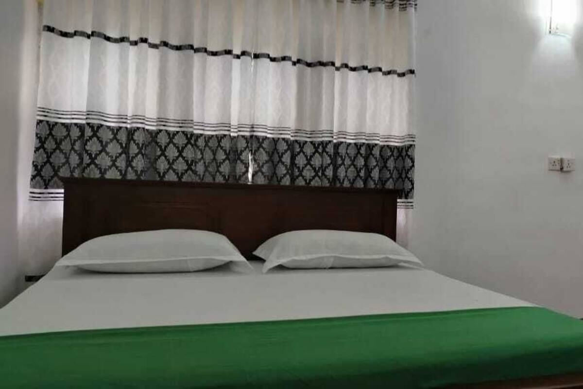 Een enkel bed met een groene sprei, witte kussens en een houten hoofdbord. Er is een raam met decoratieve gordijnen en een deur met bloemenbehang.