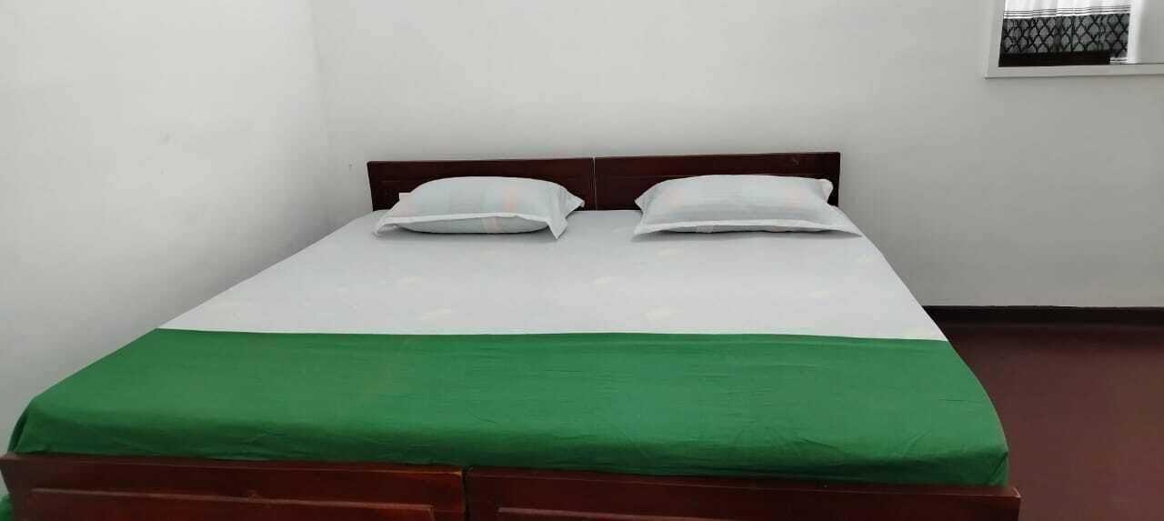 Un lit simple avec une couvre-lit verte et des oreillers blancs, dans une pièce aux murs blancs et dotée d’une fenêtre avec un store à motifs.