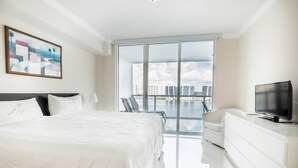 A modern hotel room with a large bed, a TV, a white dresser, and a balcony with a city view. - Le Marina Bay 1006 STR-01435 (Sunny Isles Beach)