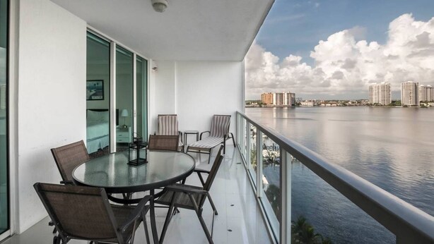 A balcony with a glass table and chairs, overlooking a calm body of water and a cityscape. - Le Marina Bay 1006 STR-01435 (Sunny Isles Beach)