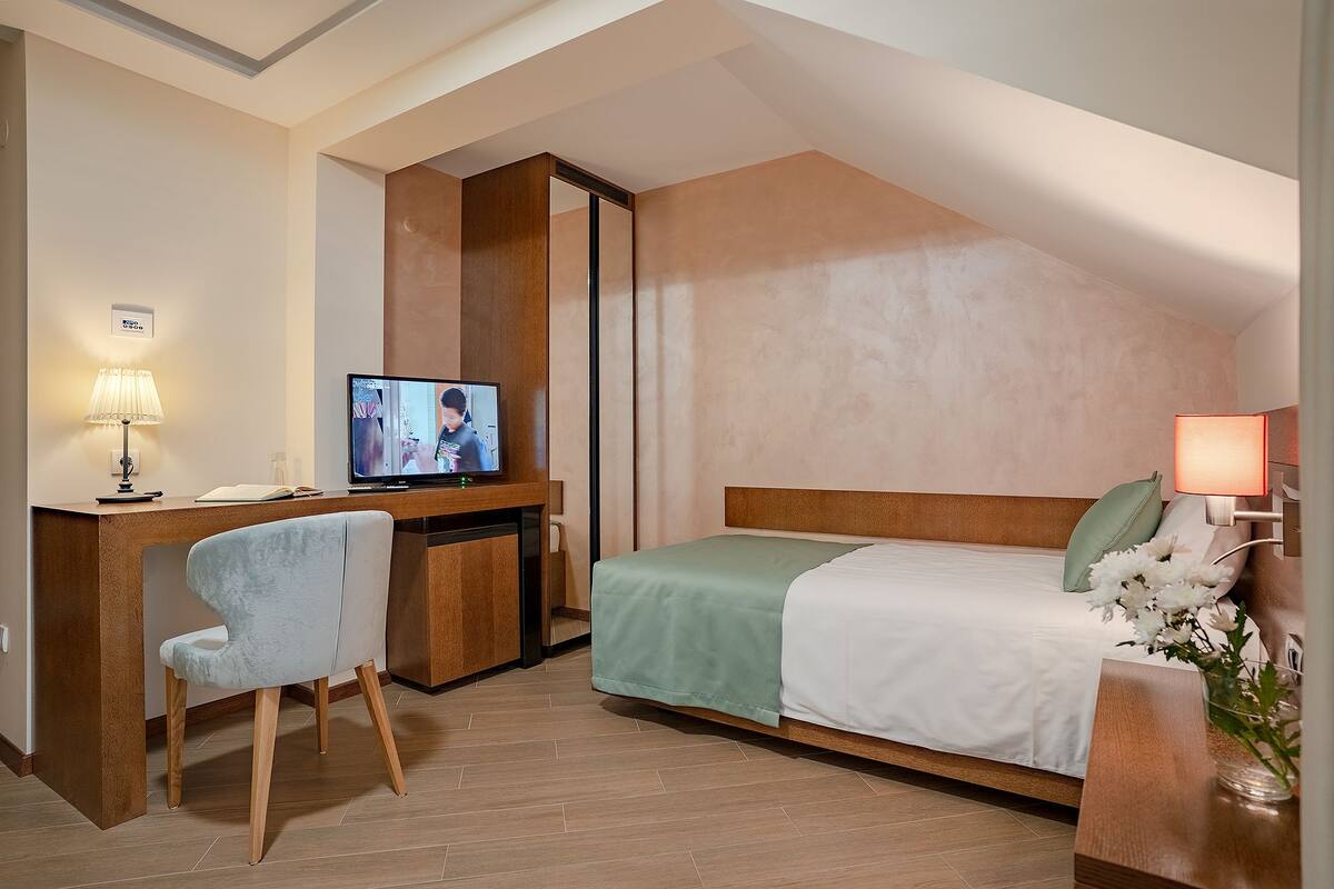 A hotel room with a bed, a desk with a television, a chair, and a bedside table with a lamp.