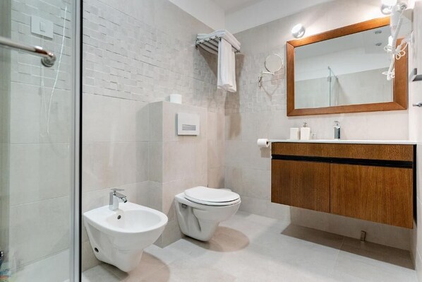 A modern bathroom with a glass shower enclosure, a white bidet, a toilet, and a wooden vanity with a mirror.