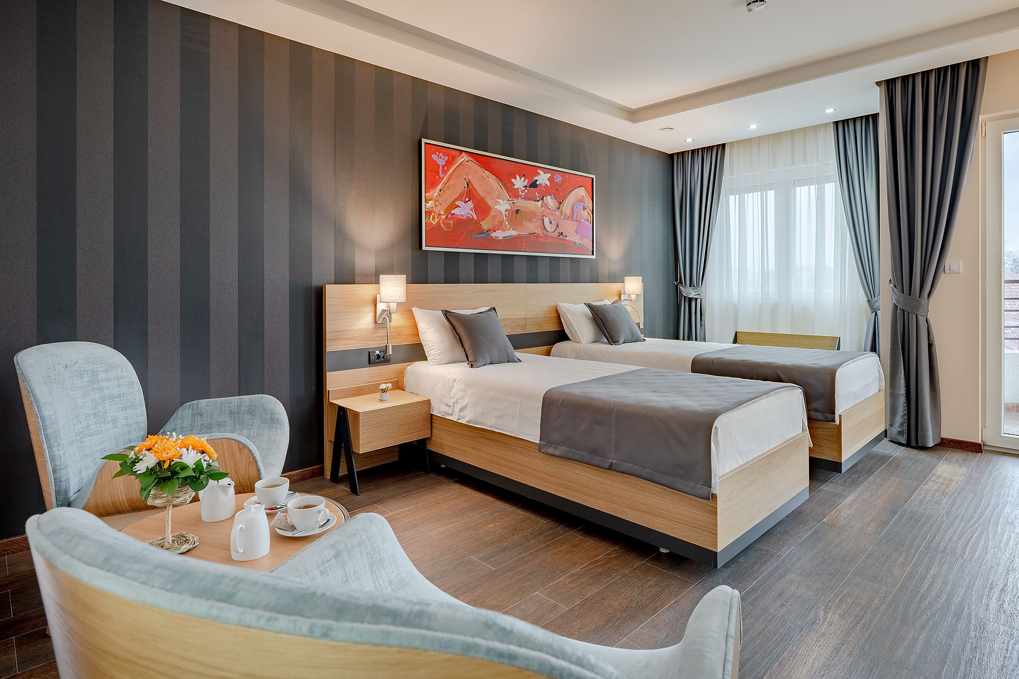 A hotel room with a large bed, a smaller bed, a seating area with a table, and a large window with curtains.