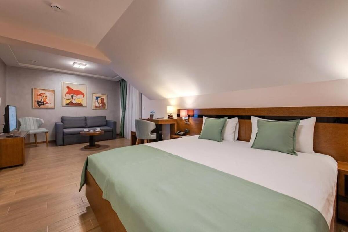 A hotel room with a large bed, a sofa, a desk, and a chair. There are paintings on the wall and a ceiling light.