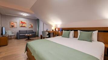Habitación de hotel con una cama grande, un sofá, un escritorio y una silla. Hay cuadros en la pared y una lámpara en el techo.