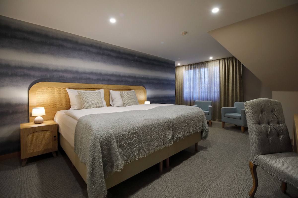 Une chambre d’hôtel moderne avec un grand lit, une table de chevet avec une lampe, une chaise et une vue sur l’extérieur par une fenêtre avec des rideaux.