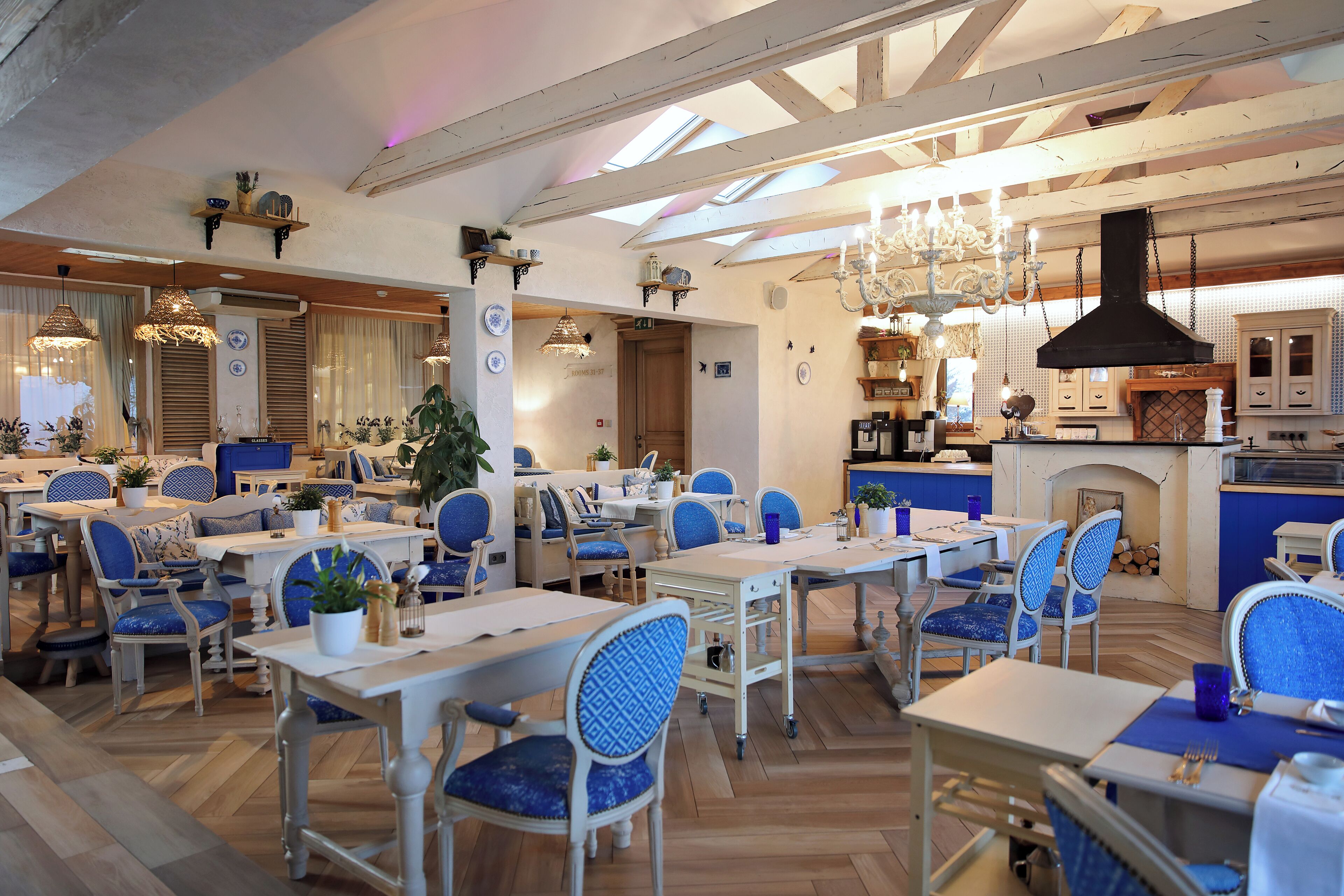 A restaurant with a high ceiling, exposed beams, and a mix of blue and white furnishings. Tables are arranged in a casual layout, and there is a fireplace in the background.
