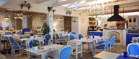 Un restaurante con techo alto, vigas a la vista y una combinación de mobiliario azul y blanco. Las mesas están dispuestas de manera informal, y se aprecia una chimenea al fondo.