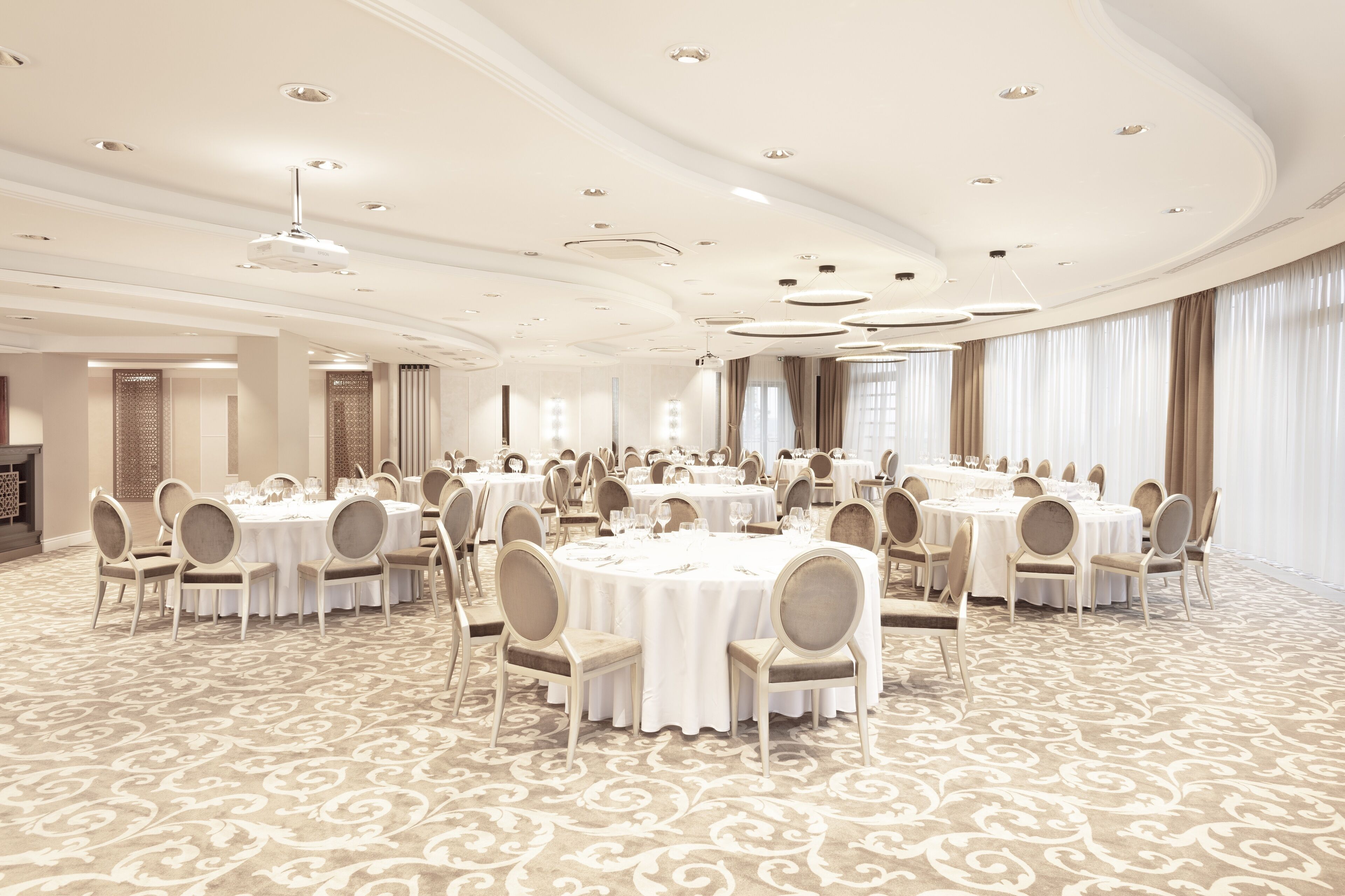 A spacious banquet hall with multiple round tables, each set with white tablecloths and chairs, and a patterned carpet.
