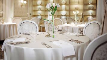 A dining area with round tables, white tablecloths, and chairs. Each table is set with cutlery, wine glasses, and a centerpiece of flowers.