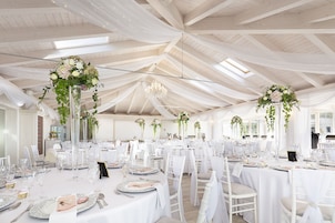 Un salón de banquetes con mesas redondas, sillas blancas y centros de mesa florales.