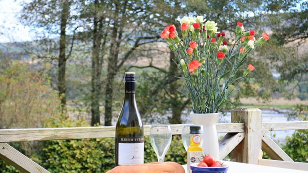A table with a bottle of wine, a glass, a baguette, cheese, a bowl of tomatoes, and a vase of flowers. - Shelduck Cottage, shoreline location, pet friendly, sleeps 7 (Glendaruel)