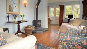 A cozy living room with a fireplace, a comfortable sofa, a television, and a dining table with a lamp. - Shelduck Cottage, shoreline location, pet friendly, sleeps 7 (Glendaruel)