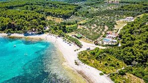Vue aérienne d’une zone côtière avec une plage, des eaux turquoise et limpides, et une zone forestière dense.