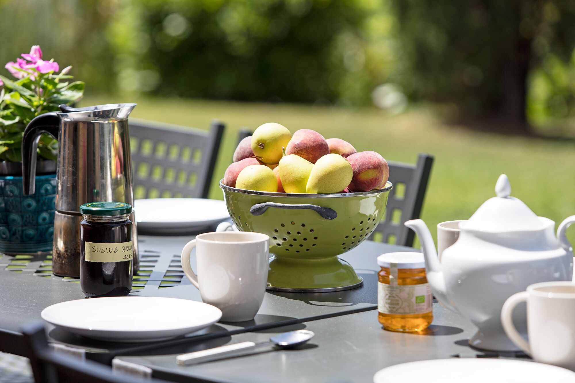 A table set for breakfast with a colander of fruit, a coffee pot, a jar of honey, and a teapot.
