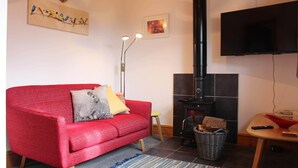 A living room with a red sofa, a fireplace, a flat-screen TV, and artwork on the walls. - Delightful Detached Cottage close to Exeter, Dartmoor and the coast. (Longdown)