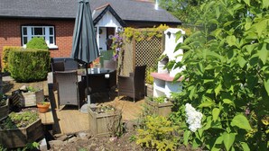 A garden with a patio area, including a table and chairs, surrounded by plants and a wooden fence. - Delightful Detached Cottage close to Exeter, Dartmoor and the coast. (Longdown)