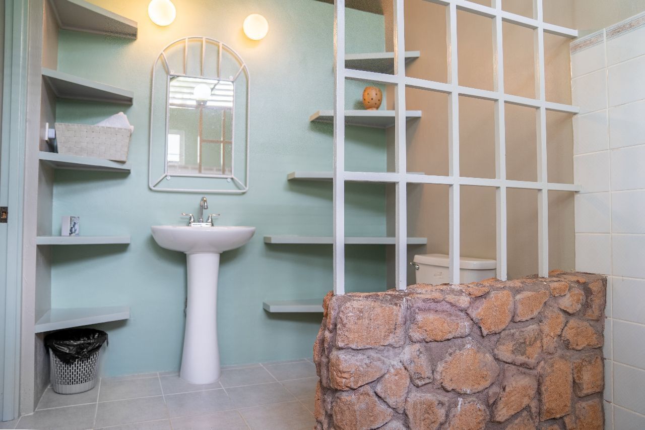 Un bagno con un lavandino bianco, uno specchio, mensole, una parete decorata in pietra e pavimento piastrellato.