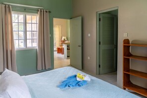 A bedroom with a bed, a wooden shelf, and a view of a dining area through an open door. - Kootney Resorts (Portsmouth)