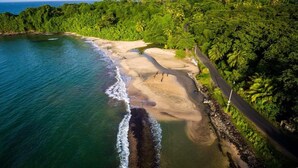 Vista aérea de una playa con aguas turquesas cristalinas, una orilla de arena y vegetación tropical exuberante.