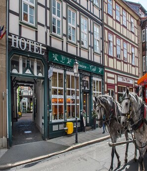Un hotel tradizionale europeo con un cartello ben visibile che riporta la scritta 'HOTEL', un tendone verde e una carrozza trainata da cavalli sulla strada.