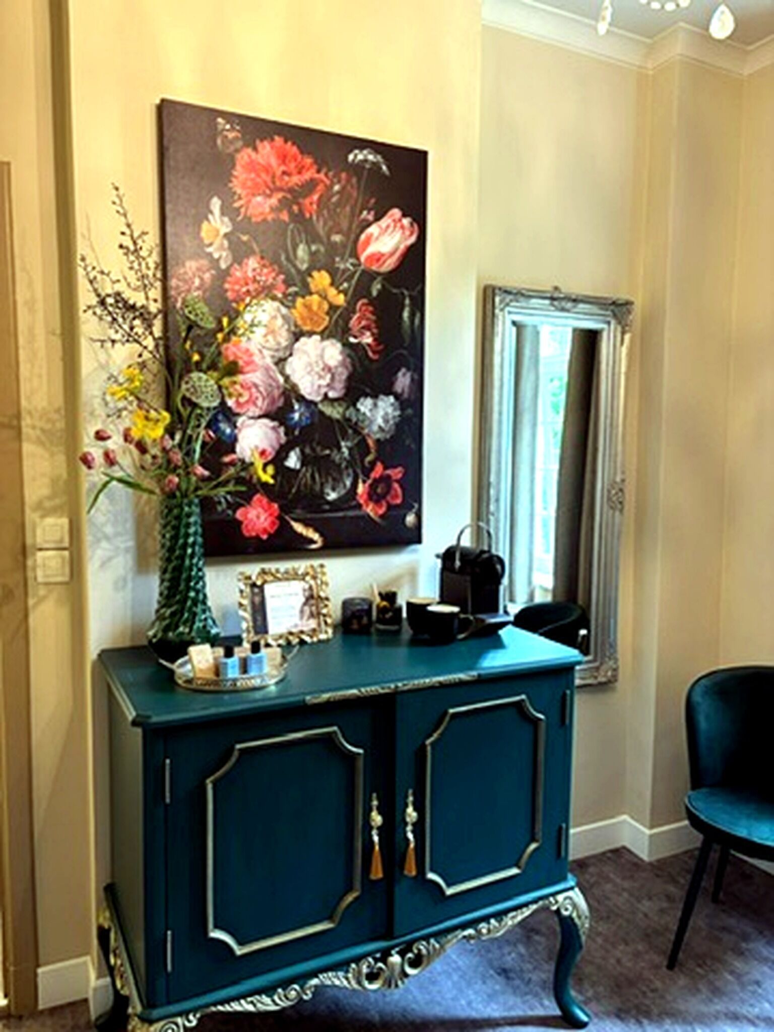 Una consola color turquesa con un cuadro floral encima, un espejo y una silla.