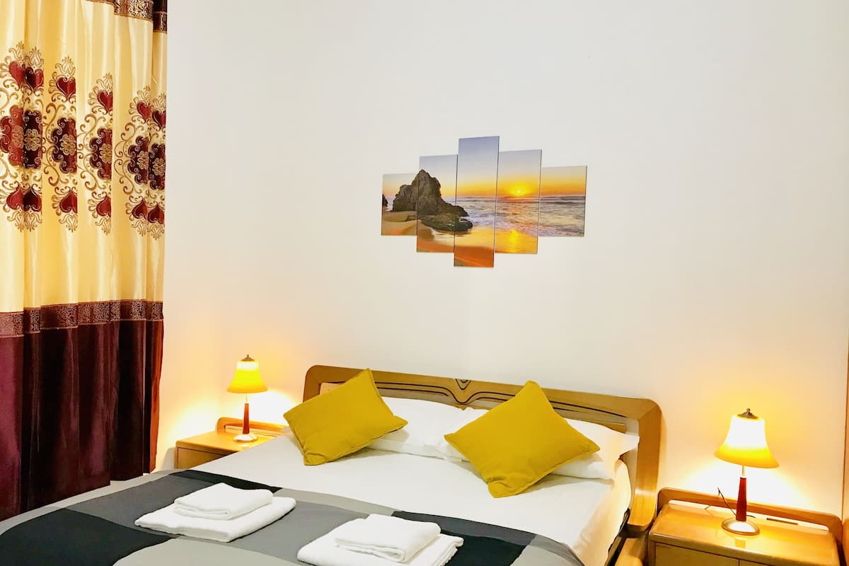 A neatly made bed with two pillows, a bedside table with a lamp, and a wall-mounted artwork depicting a sunset over a rocky landscape.