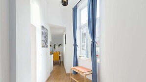 Un pasillo estrecho con un banco de madera, una ventana con cortinas azules y una silla amarilla al fondo.