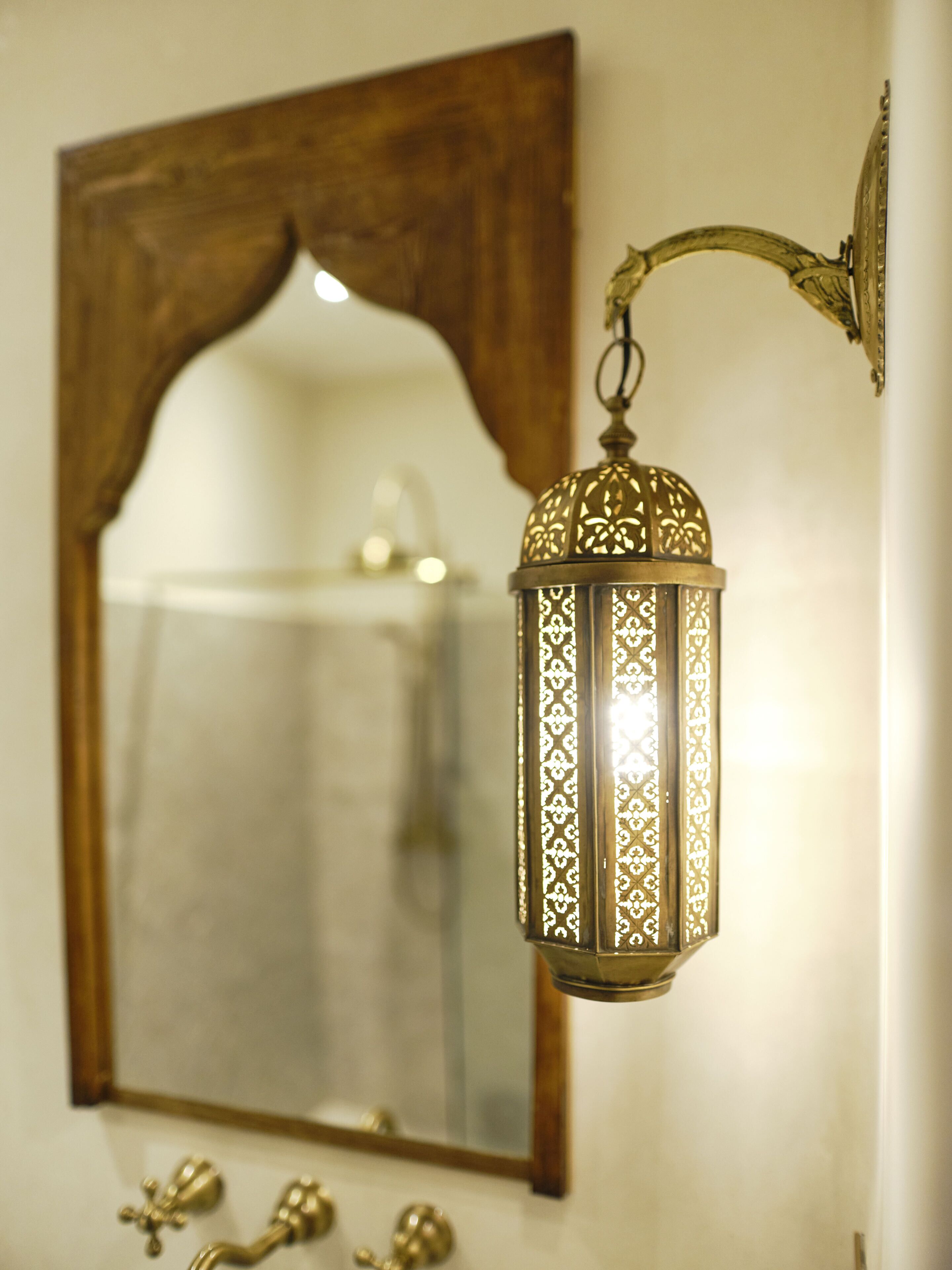 A wall-mounted lantern with intricate patterns, a wooden-framed mirror, and brass fixtures below.