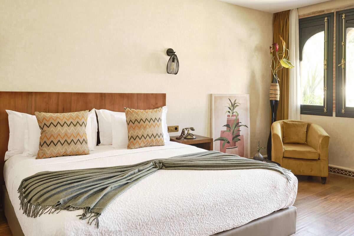 A hotel room with a large bed, a wooden headboard, a nightstand with a telephone, a chair, and a window with curtains.