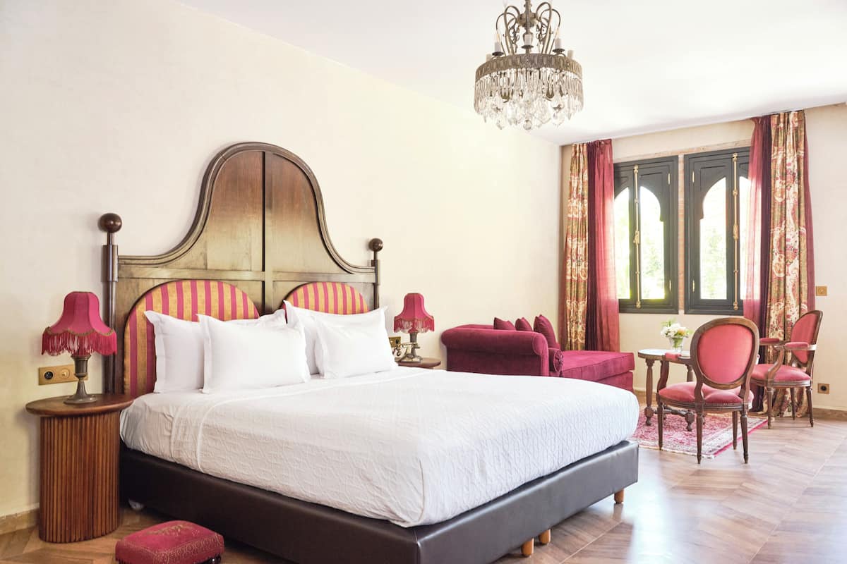 A large bed with a wooden headboard, two bedside tables with lamps, a red upholstered chair, a small table with a vase, and a chandelier.