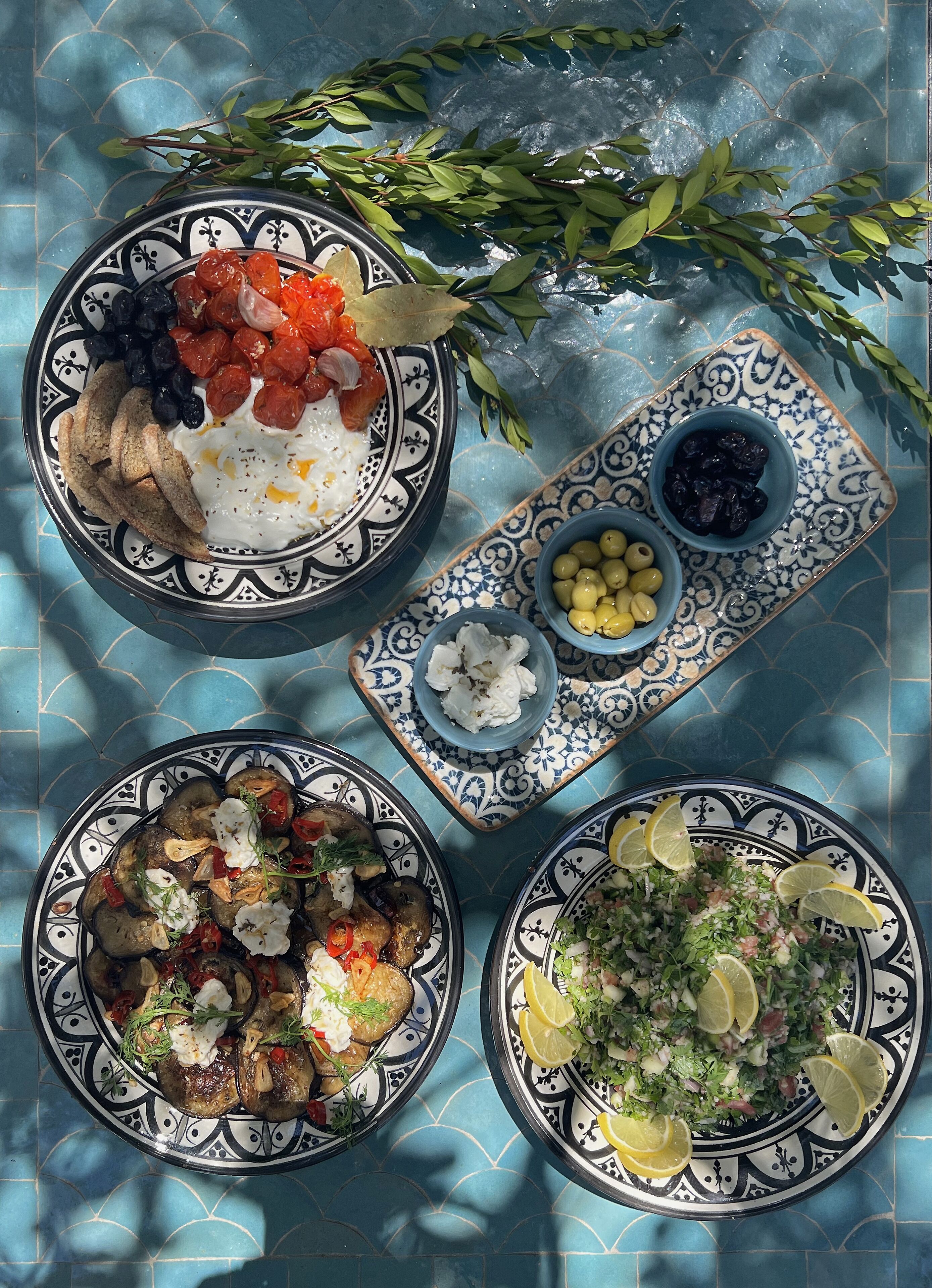Een tafel met vier borden eten, waaronder een salade, een gerecht met eieren en tomaten, een bord met olijven, een schaal olijven, en een bord met gegrilde groenten en feta.