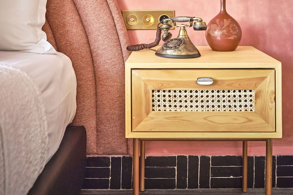 A bedroom with a wooden nightstand, a vintage telephone, a vase with a plant, and a decorative wall piece.
