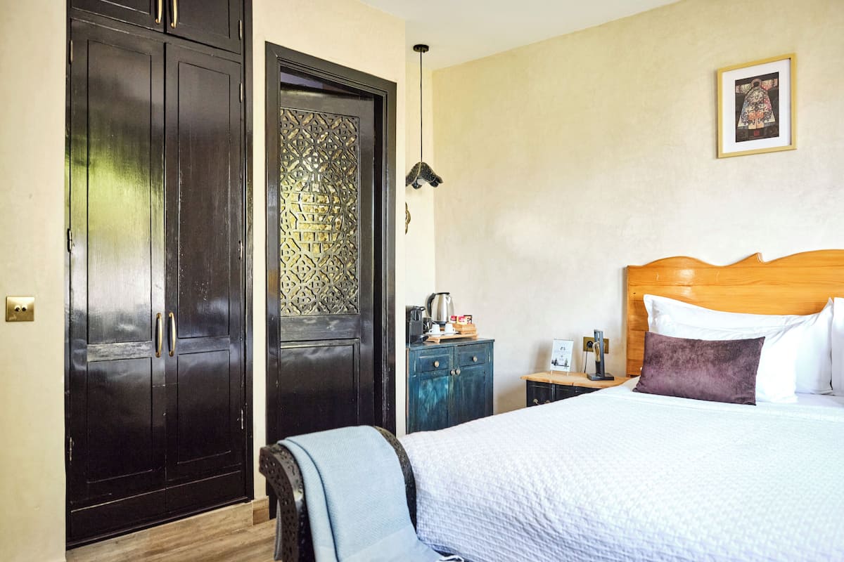 A hotel room with a large bed, a wooden headboard, a nightstand with a lamp, a wardrobe, and a decorative door.