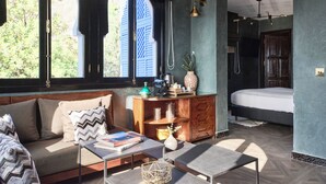 Una habitación moderna con sofá, una mesita y una cama. Se observan ventanas con contraventanas, un mueble de madera y una lámpara fijada a la pared.