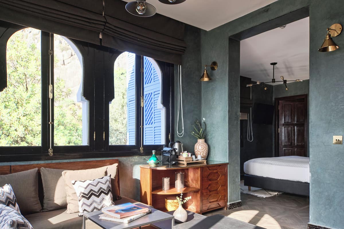 A modern room with a sofa, a small table, and a bed. There are windows with shutters, a wooden cabinet, and a wall-mounted light fixture.