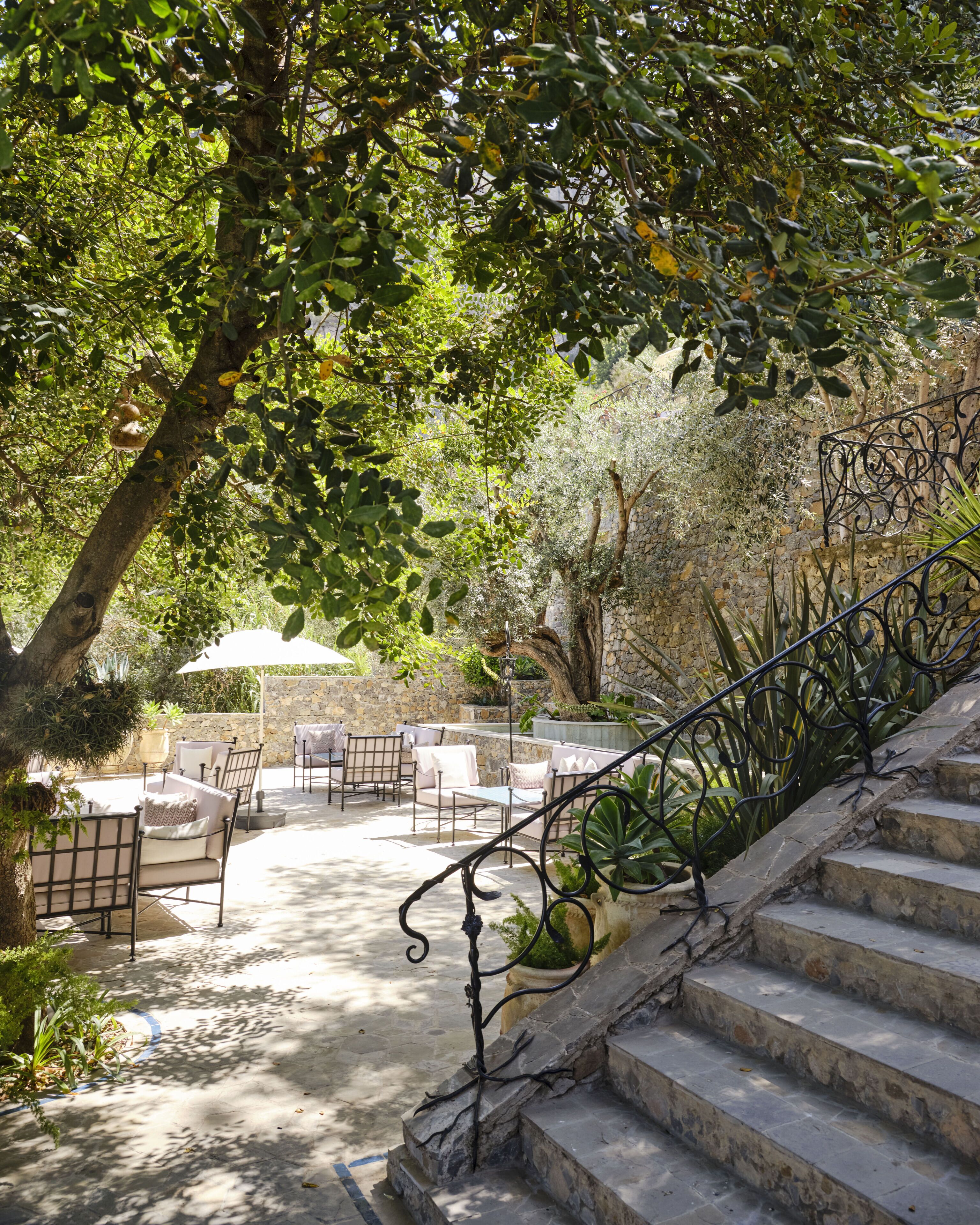 A stone staircase leading to an outdoor seating area with chairs and tables, surrounded by greenery and a stone wall.