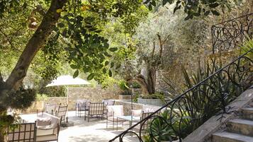 A stone staircase leading to an outdoor seating area with chairs and tables, surrounded by greenery and a stone wall.