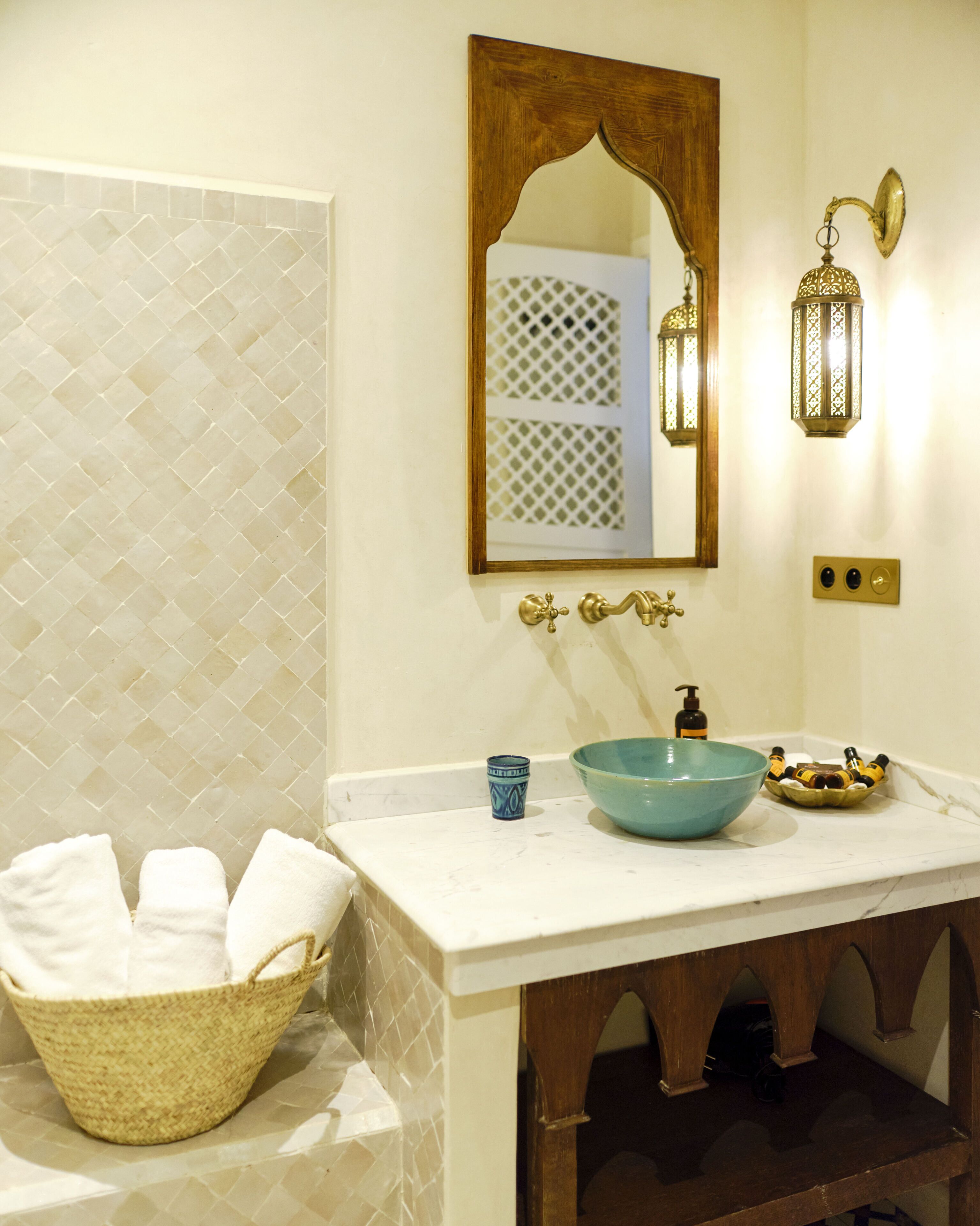 Une salle de bain avec un mur recouvert de carreaux blancs, un miroir encadré en bois, un lavabo bleu, un luminaire fixé au mur et un panier en osier contenant des serviettes.