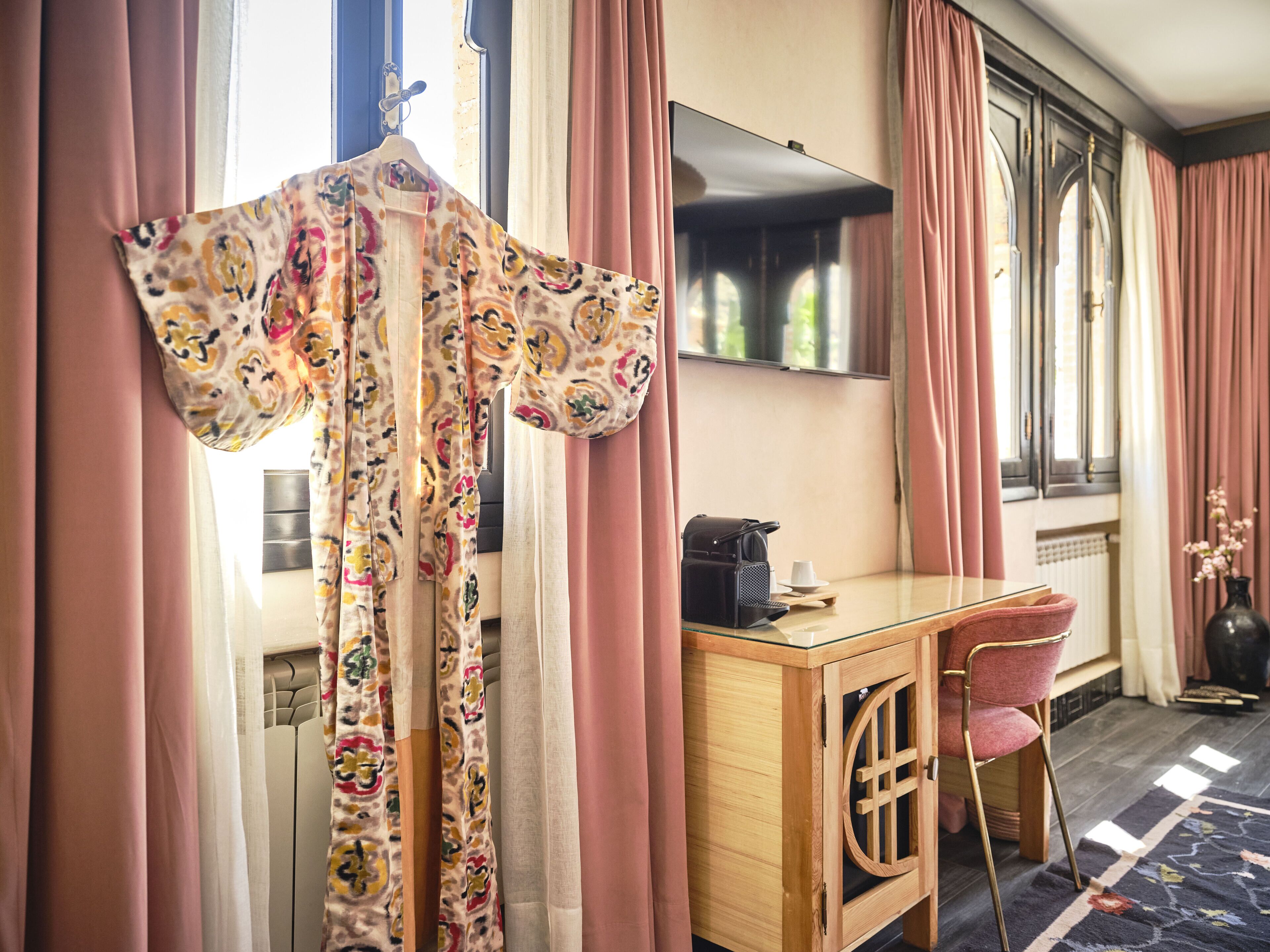Habitación de hotel con un escritorio de madera, una silla, una bata colgada y cortinas rosas.