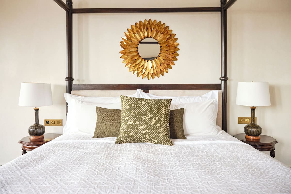 A four-poster bed with white linens and patterned pillows, a gold leaf mirror above, and two bedside tables with lamps.