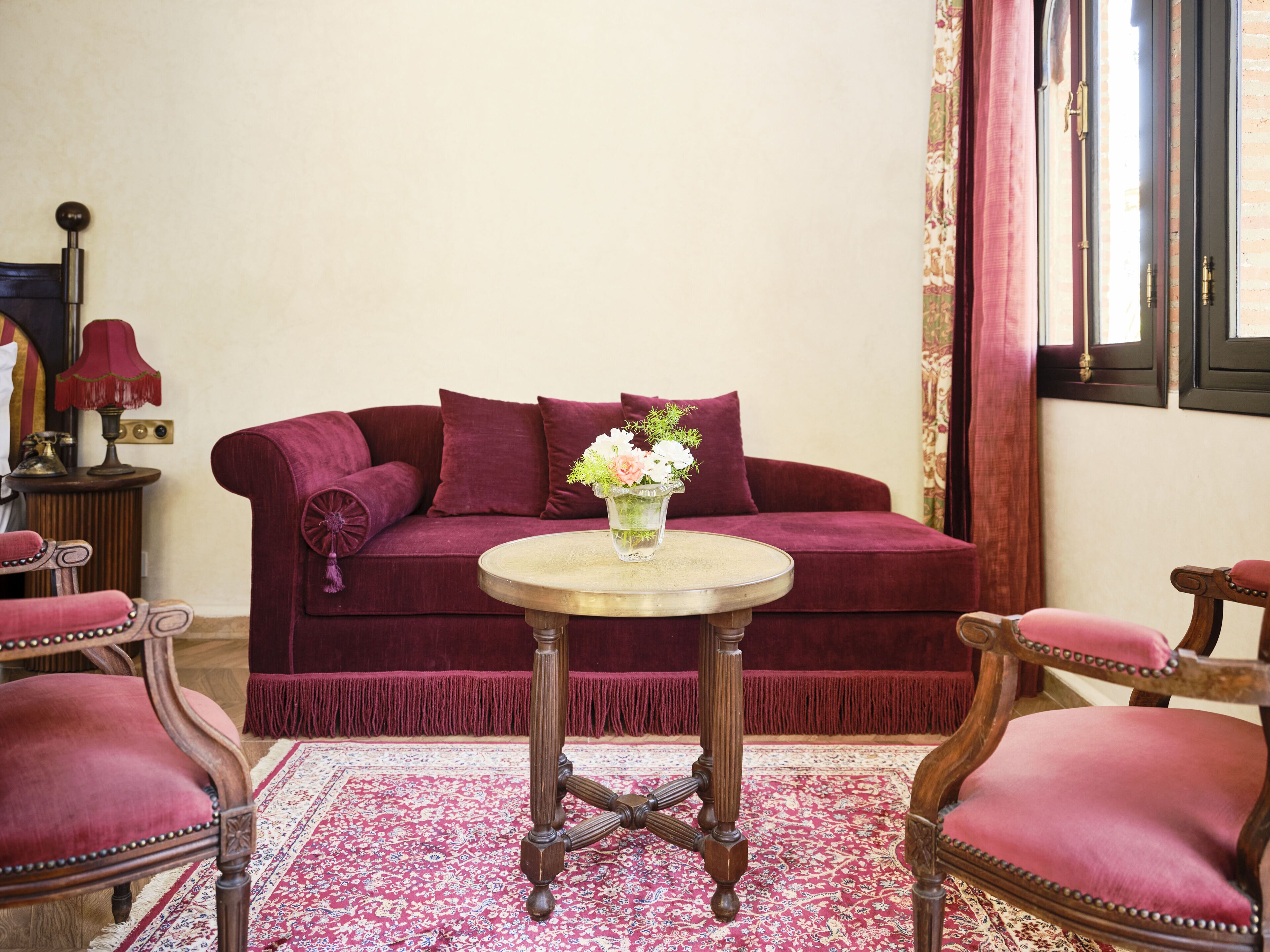 A room with a red sofa, a round wooden table with a vase of flowers, and two matching chairs.
