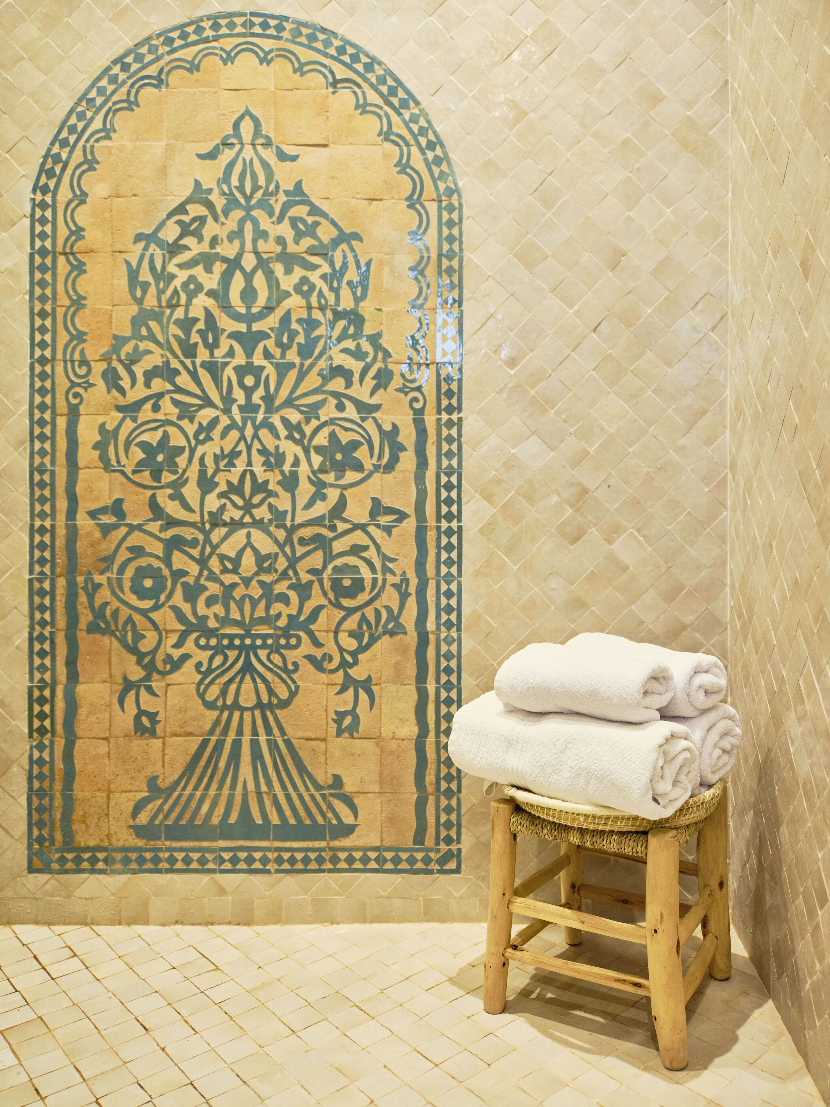 Un baño con un muro de azulejos con estampado floral, un taburete de madera con toallas blancas dobladas y piso de azulejos.